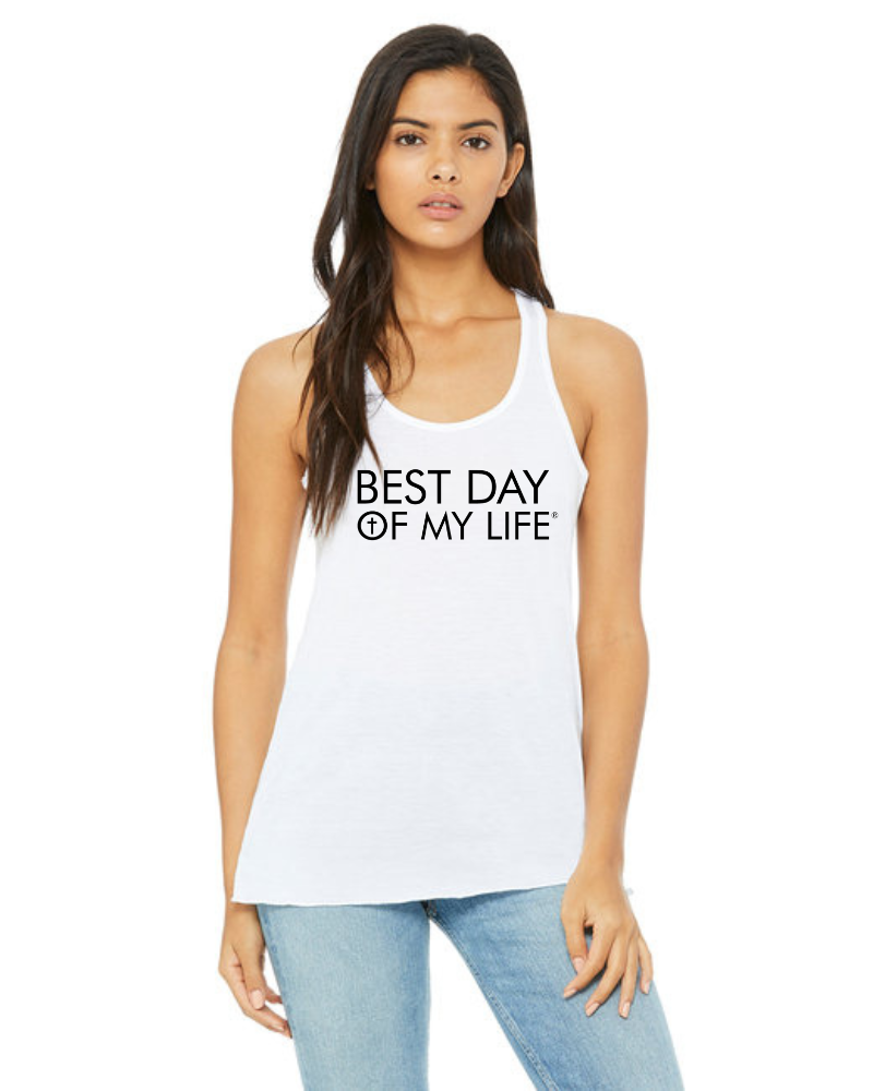 medium skinned person with long hair wearing a white tank top with best day of my life written across in black and a cross in the O