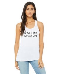 medium skinned person with long hair wearing a white tank top with best day of my life written across in black and a cross in the O