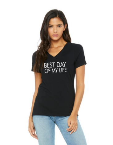 medium-skinned person with long brown hair wearing a black vneck t-shirt with best day of my life written across in white.