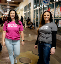 two white women smiling, one on left wearing a pink crewneck with best day of my life written across in white and jeans, one on right wearing a grey crewneck t-shirt with best day of my life written in white over a black long-sleeved shirt