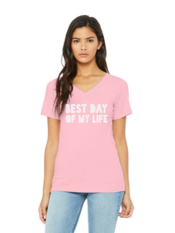 medium-skinned person with long brown hair wearing a light pink vneck t-shirt with best day of my life written across in white block lettering.