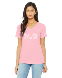 medium-skinned person with long brown hair wearing a light pink vneck t-shirt with best day of my life written across in white with a cross in the O.