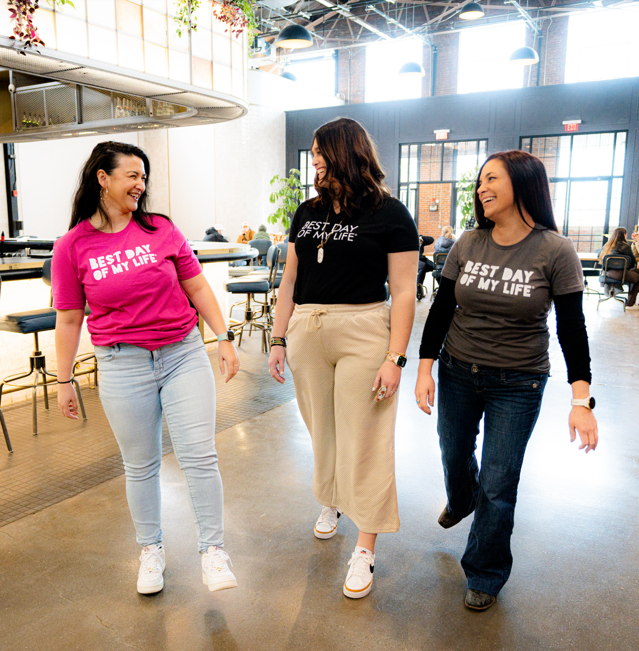 three light-skinned people with medium to long brown hair looking at each other and laughing. on the left the person is wearing a pink crewneck t-shirt with best day of my life written across in white block lettering, in the center the person is wearing a black v-neck with best day of my life written across in white, on the right the person is wearing a grey crewneck t-shirt with best day of my life written in block lettering over a long-sleeved black shirt.