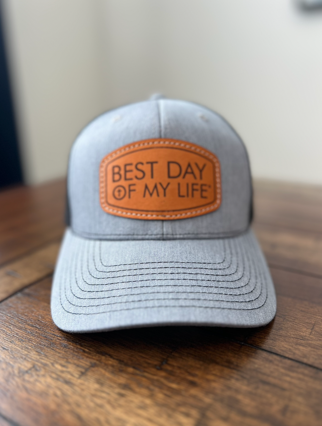Gray cap with a brown leather patch reading 'Best Day of My Life' on a wooden surface.
