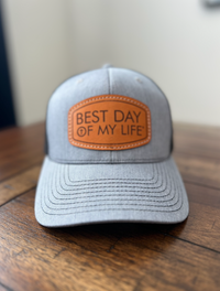 Gray cap with a brown leather patch reading 'Best Day of My Life' on a wooden surface.