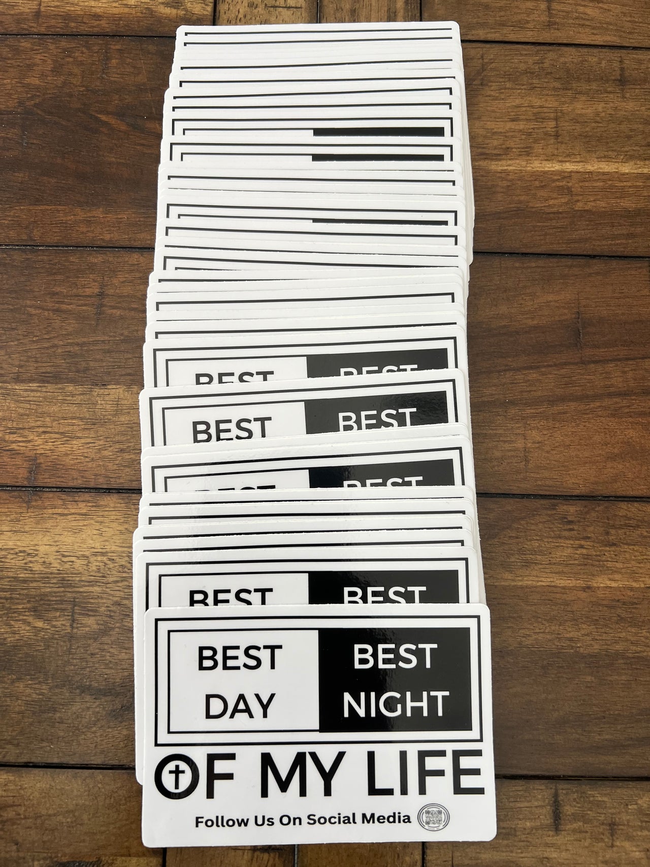 stack of stickers on wooden background, graphic of a white and black sticker on a grey background. sticker reads best day best night of my life with a cross