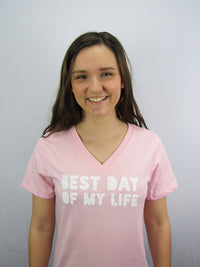 Thumbnail for half body of a light-skinned person with long brown hair smiling wearing a light pink vneck t-shirt with best day of my life written in white block lettering. 
